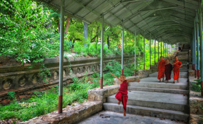 Young monks walking and playing along a covered pathway surrounded by greenery in Myanmar.