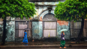 Burmese Ladies walking under umbrellas during Myanmar's monsoon season, with vibrant green trees and a weathered historic building as the backdrop