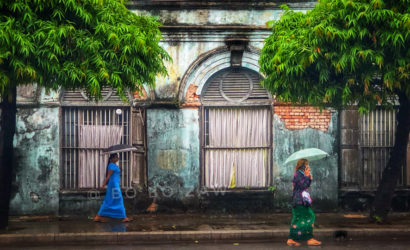 Burmese Ladies walking under umbrellas during Myanmar's monsoon season, with vibrant green trees and a weathered historic building as the backdrop