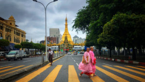 Buddhist nuns in pink robes walking across a vibrant street in Yangon with the iconic Sule Pagoda in the background, ideal for a private Yangon photo tour or Yangon photography tour highlighting the city's cultural and architectural charm