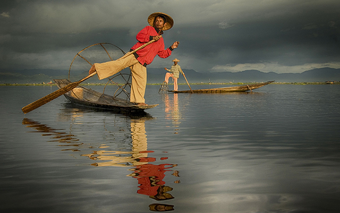 Traditional Inle fishermen balancing on boats with fishing nets, reflecting on calm waters under dramatic monsoon skies in Myanmar.