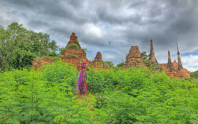 A local woman in traditional attire walking through lush green vegetation near ancient stupas under dramatic monsoon skies in Inle, Myanmar.