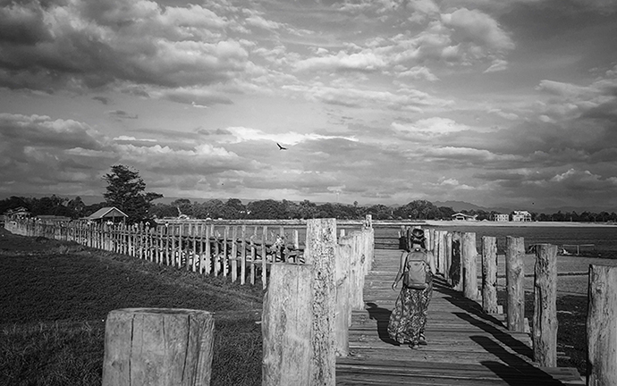 A woman walking across the historic U Bein Bridge in Mandalay, Myanmar, under a cloudy sky in a timeless black-and-white scene.