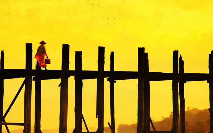 A Buddhist Nun walking across the iconic U Bein Bridge in Mandalay, Myanmar, silhouetted against a golden sunrise.