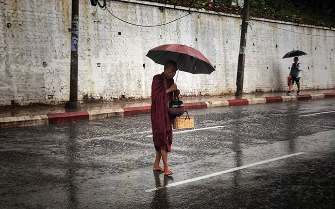 A Buddhist monk walking barefoot under an umbrella during heavy rain on a street in Yangon, Myanmar.